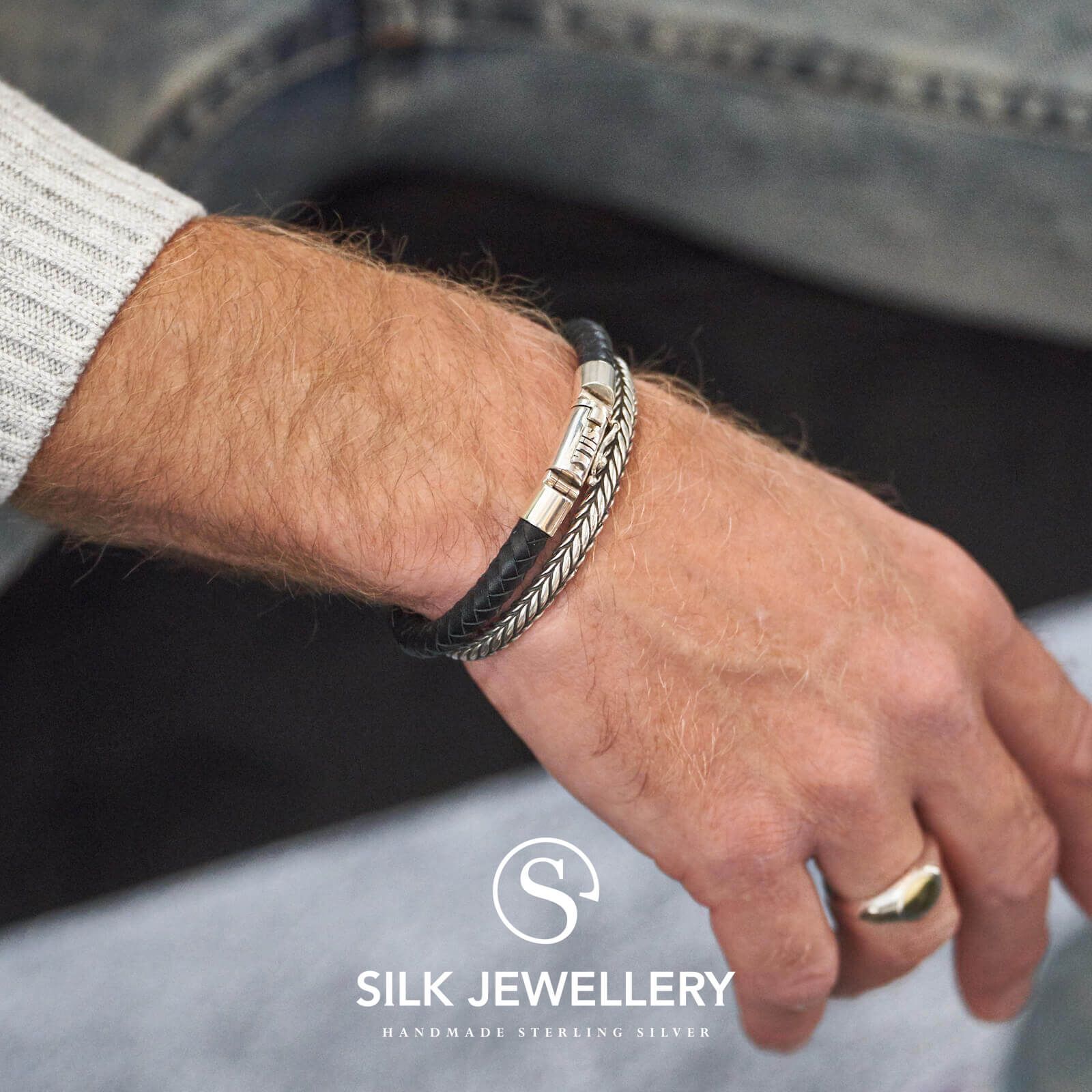 Herenpols met zilveren en zwarte leren armbanden en gouden ring, logo van Silk Jewellery zichtbaar.