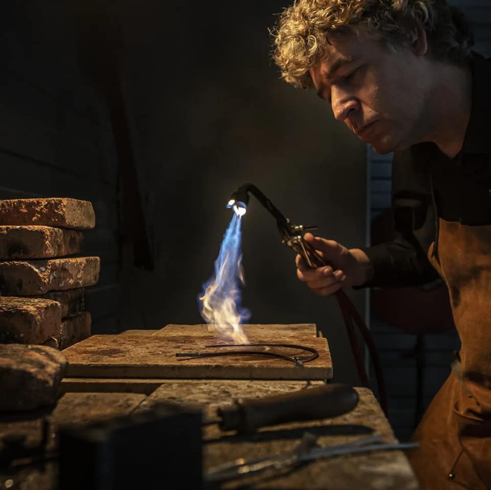 Man gebruikt een brander om een ​​stuk metaal te verhitten in een werkplaats. Blauwe vlammen, bruin schort en een stapel bakstenen.