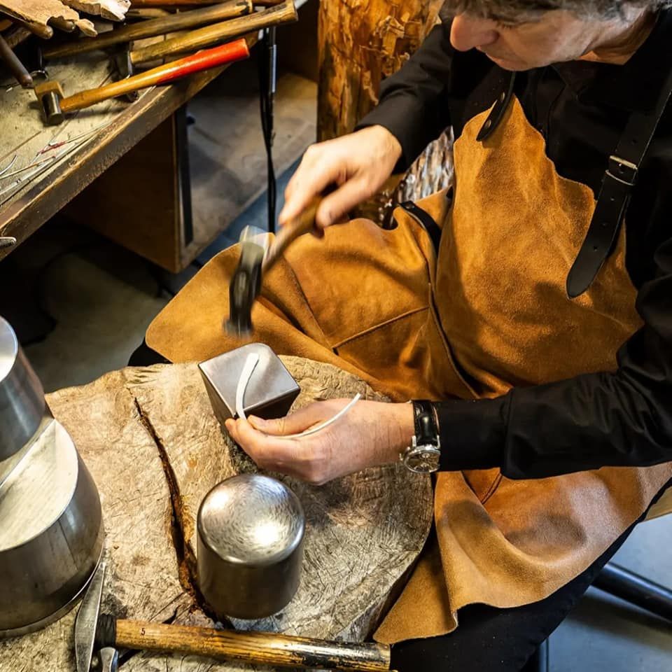 Juwelier hamert op een stuk metaal in zijn werkplaats, draagt ​​een schort en is omringd door gereedschap.