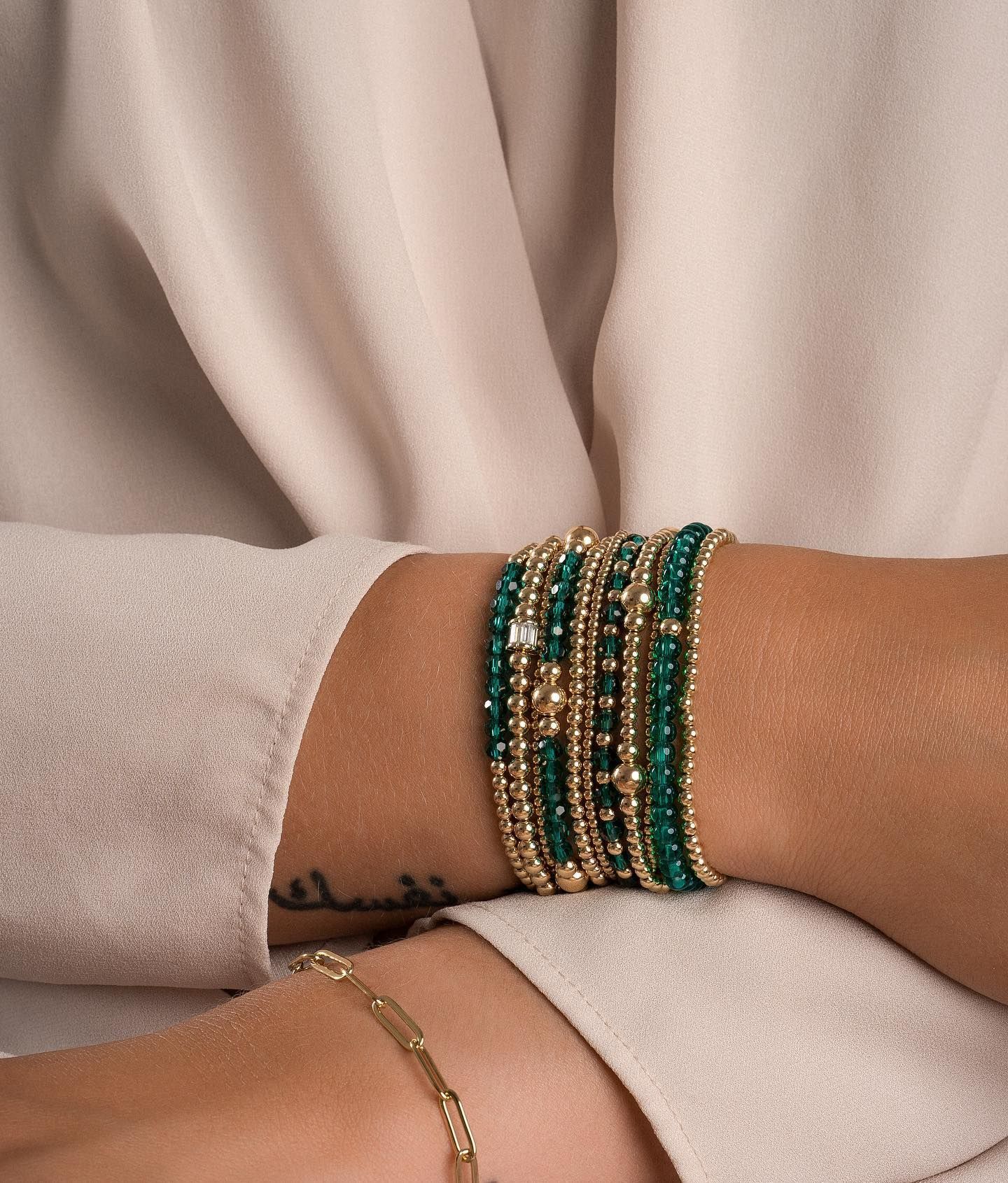 Armbanden van goud en groene kralen op elkaar gestapeld om een pols, tegen een beige top.
