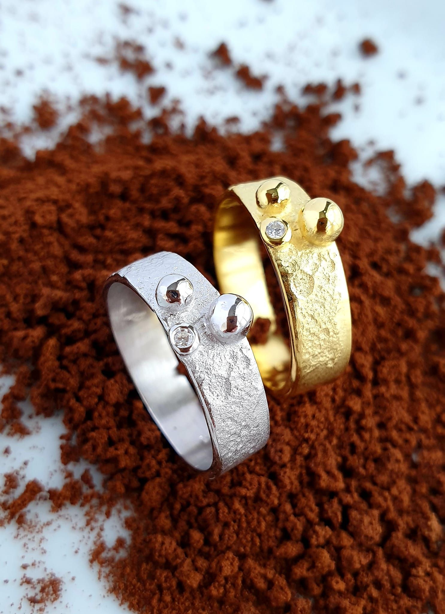 Twee ringen, één van zilver en één van goud, rusten op een bedje van bruine korrels.