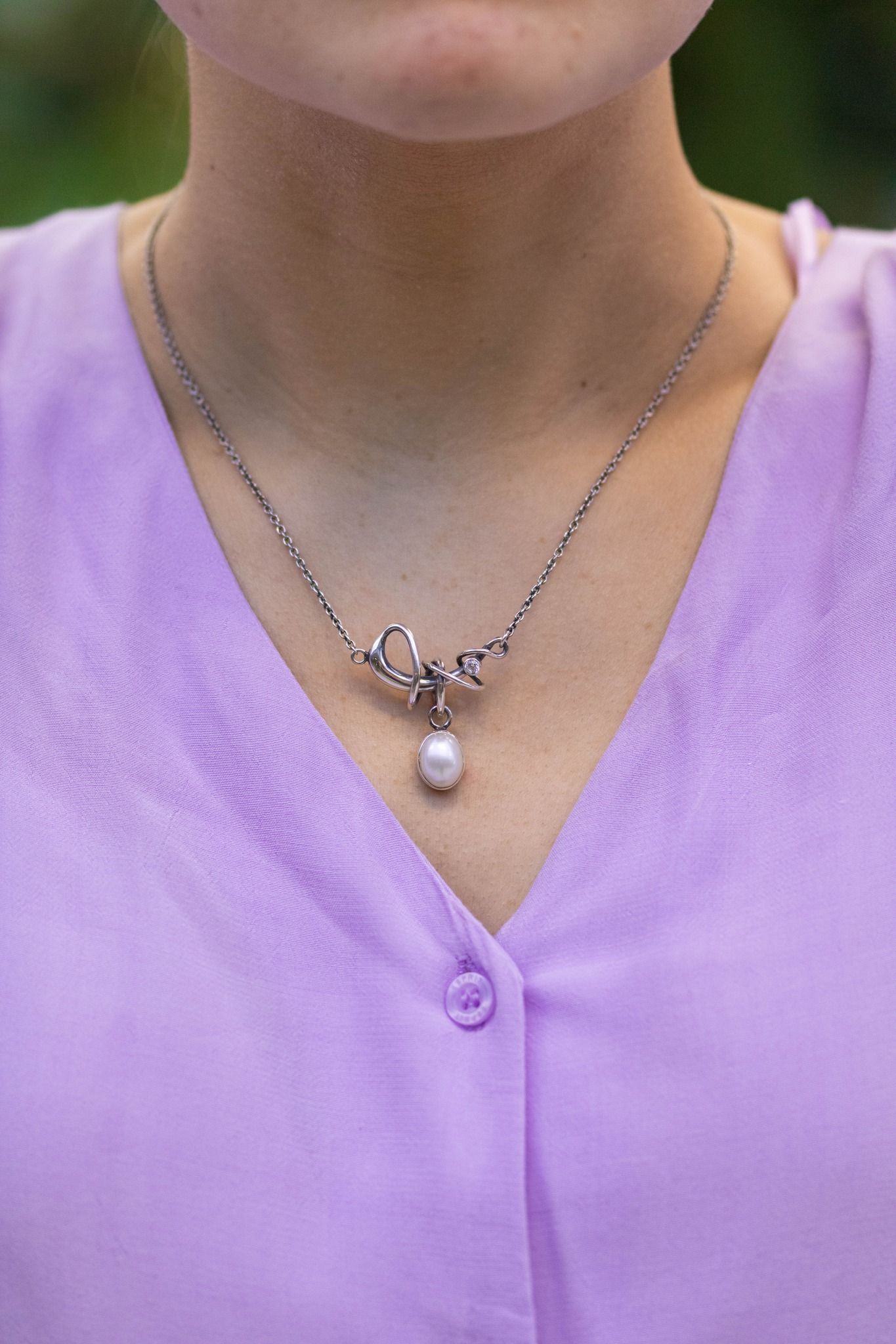 Vrouw draagt een zilveren ketting met een parelhanger en een lavendelkleurig shirt.