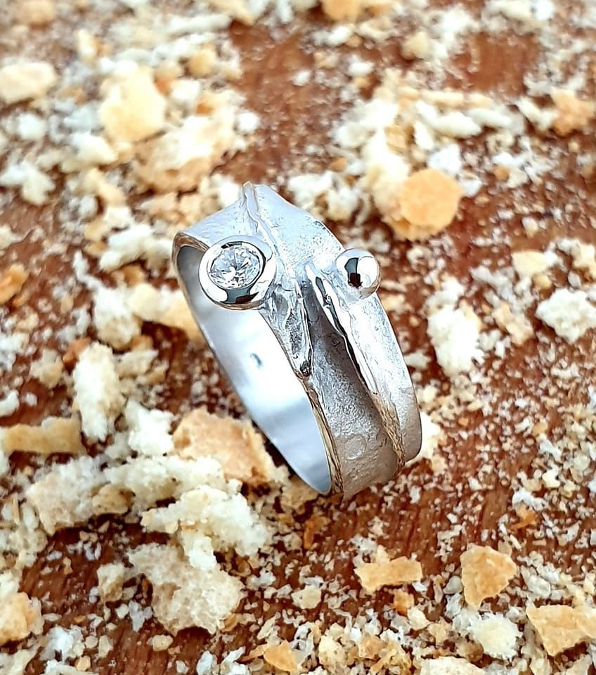 Zilveren ring met twee stenen op een gestructureerd oppervlak, rustend op een bruin oppervlak bezaaid met kruimels.