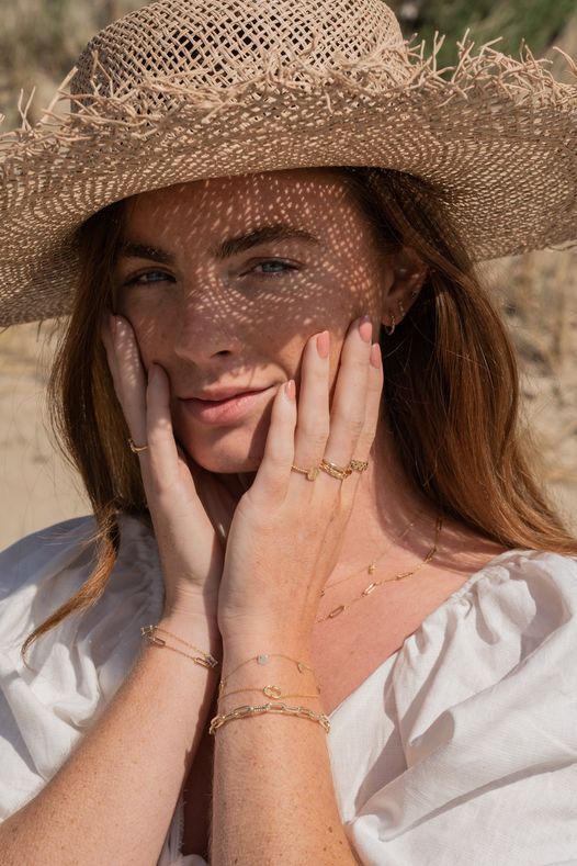 Vrouw met een strohoed, schaduw op haar gezicht, draagt gouden sieraden en raakt haar gezicht aan.
