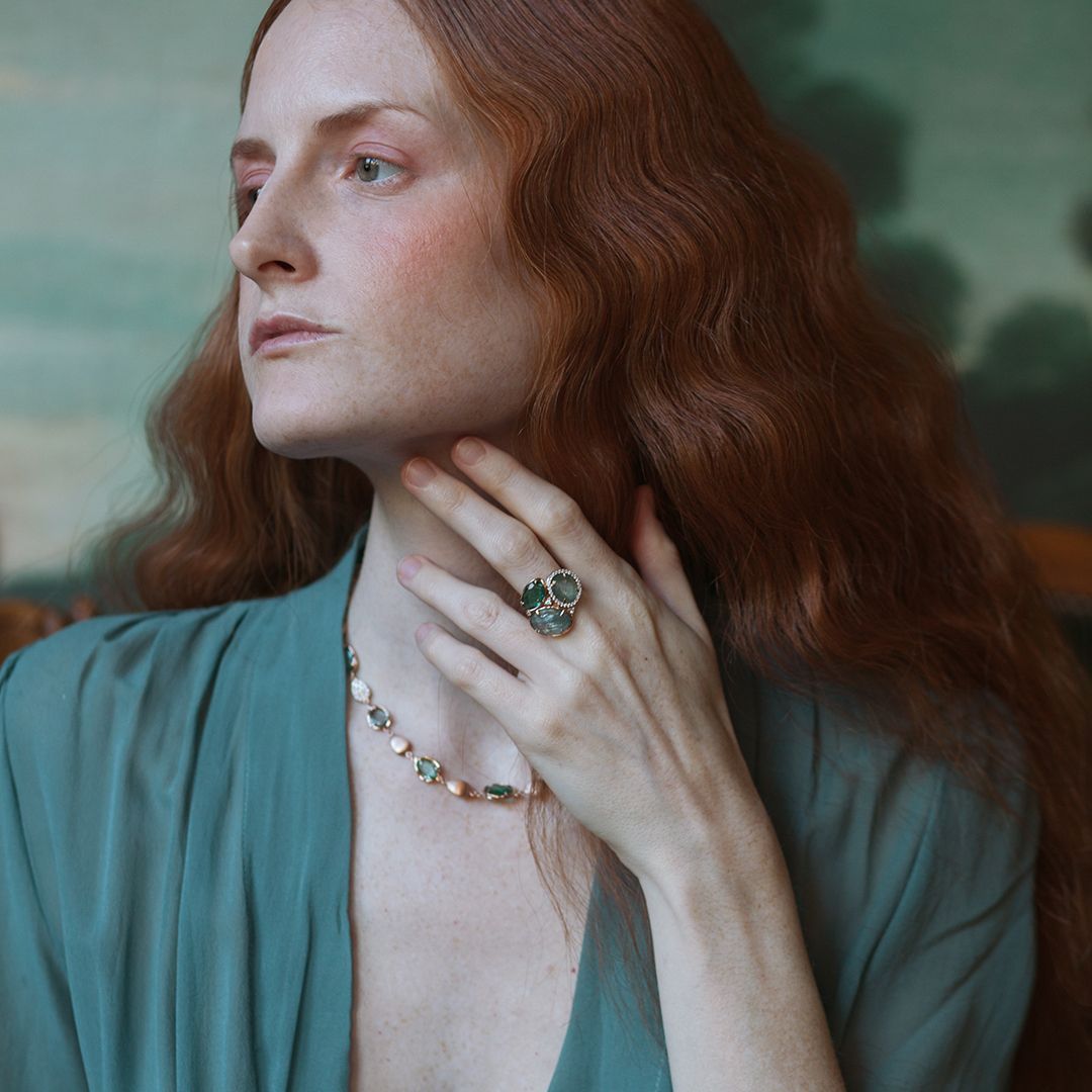 Een vrouw met rood haar draagt ​​sieraden die haar nek raken. Ze draagt ​​een blauwgroene top en staat voor een schilderij.