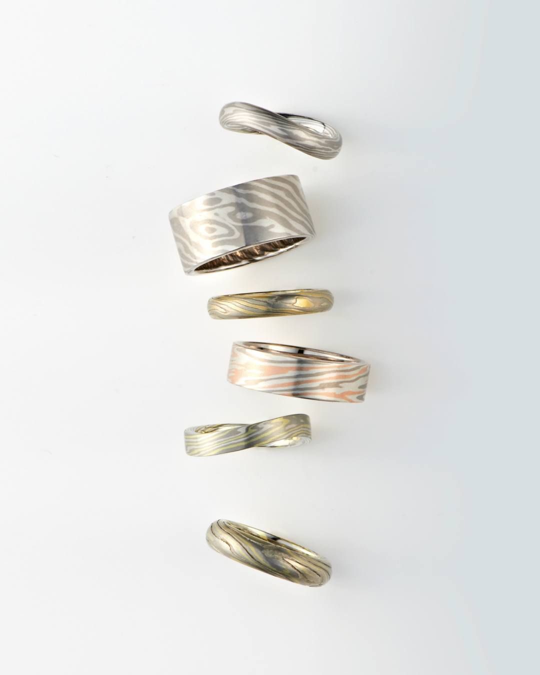 Zes metalen ringen met verschillende ontwerpen en kleuren, verticaal afgebeeld op een witte achtergrond.