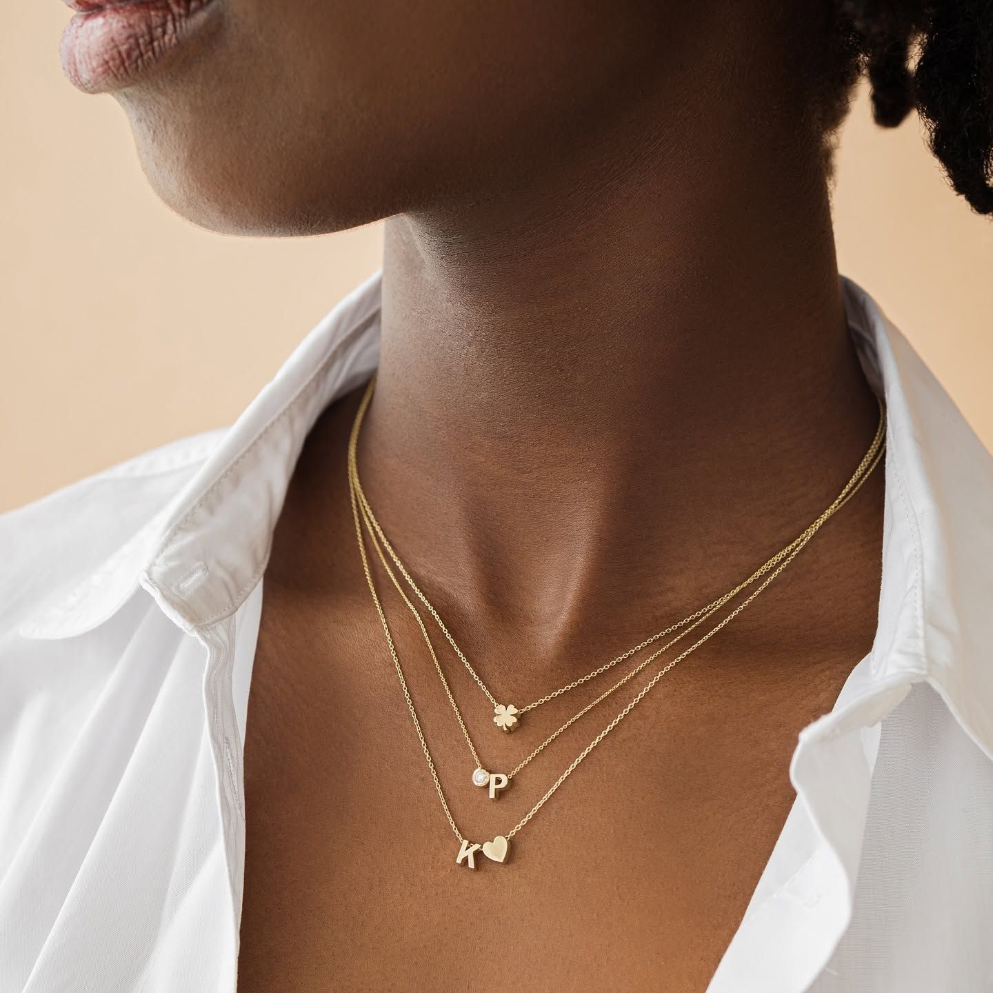 Vrouw draagt ​​een gouden ketting met hanger, een witte trui en een bruine jas.
