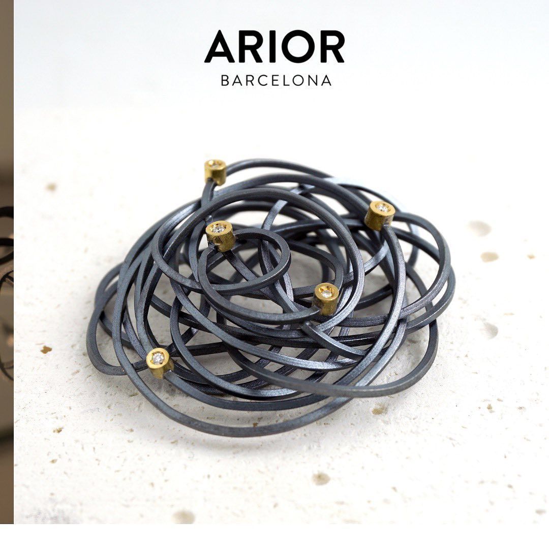Broche van donkere metalen spiralen met kleine gouden accenten, op een witte achtergrond; logo