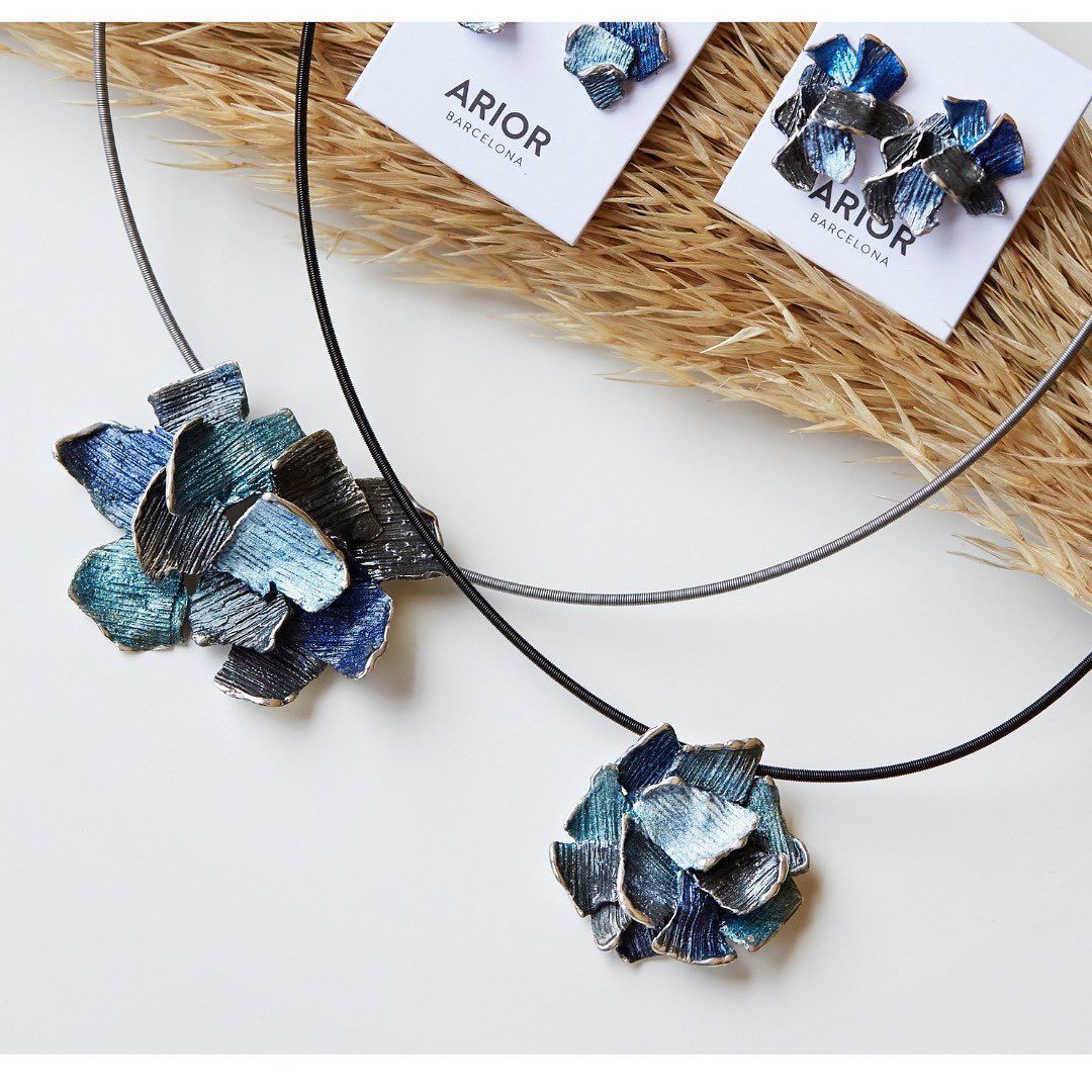 Sieradenset: ketting met bloemenhanger, bijpassende oorbellen in blauw en zilver, op een witte ondergrond.