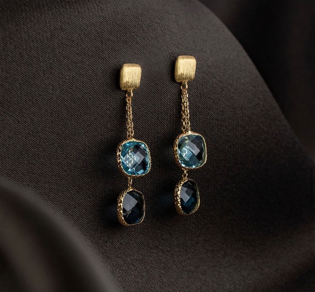 Gouden oorbellen met blauwe edelstenen op een donkere stof.