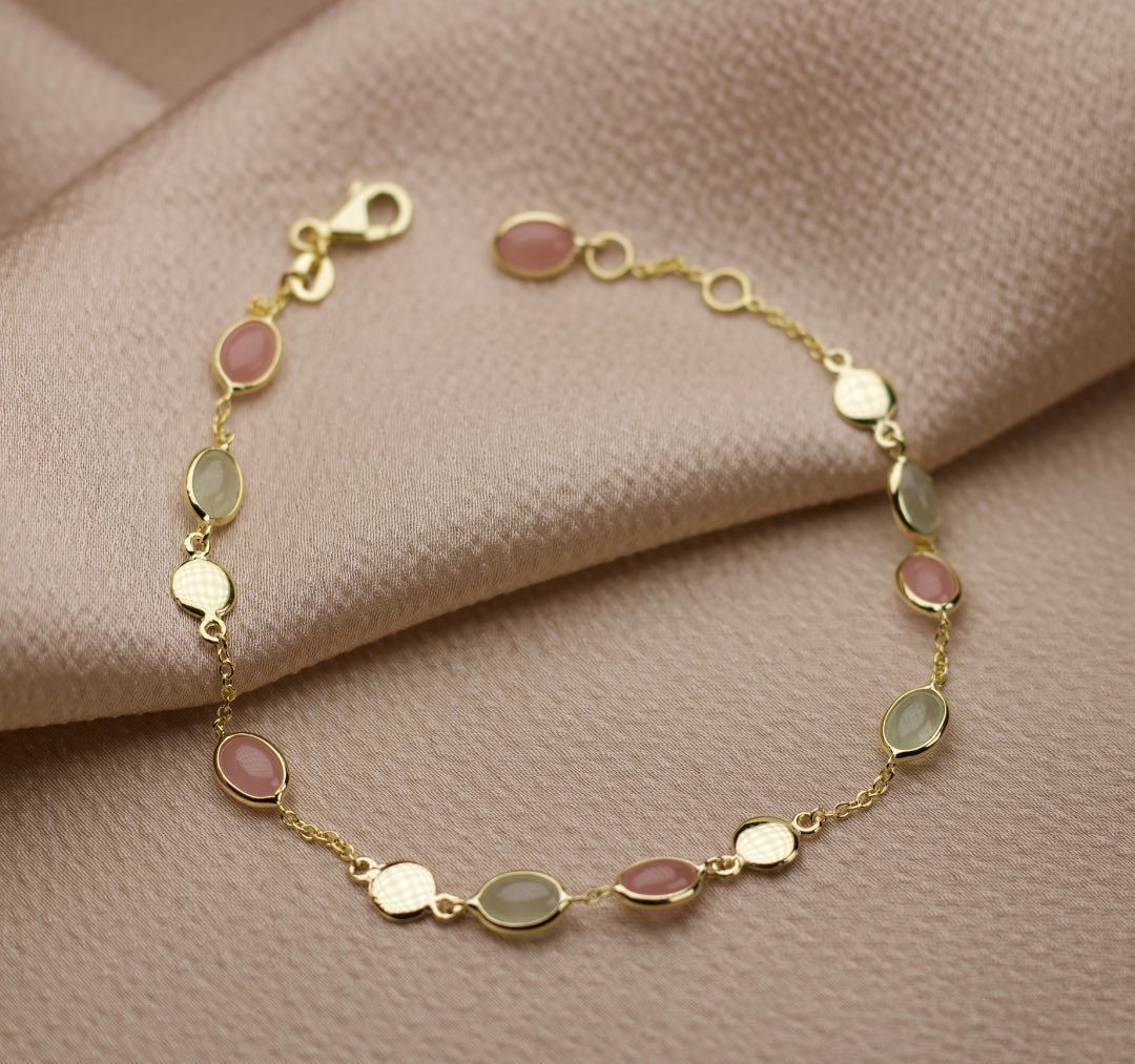 Gouden armband met ovale roze en groene stenen op een zijdeachtig oppervlak.