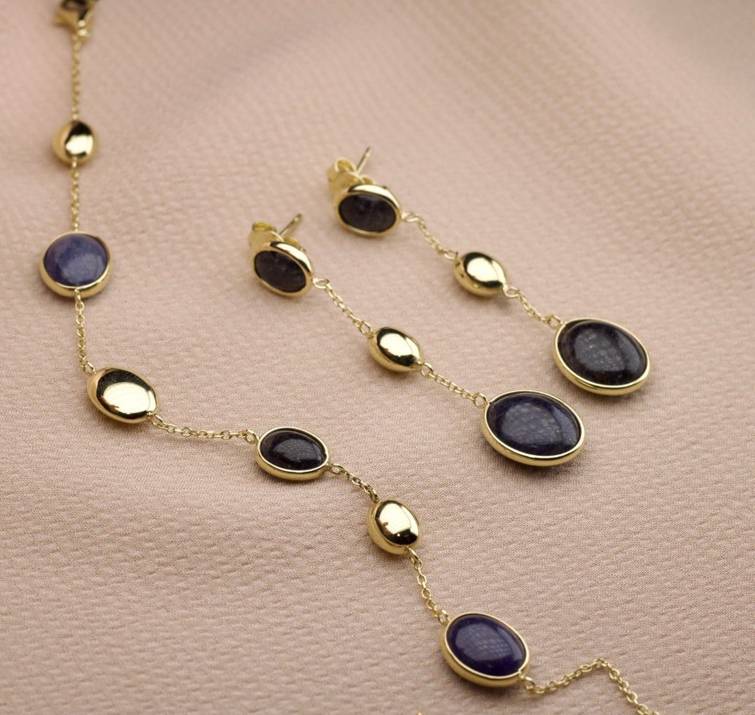 Gouden ketting en oorbellen bezet met ovale, blauwe edelstenen en gouden accenten op een crèmekleurige achtergrond.