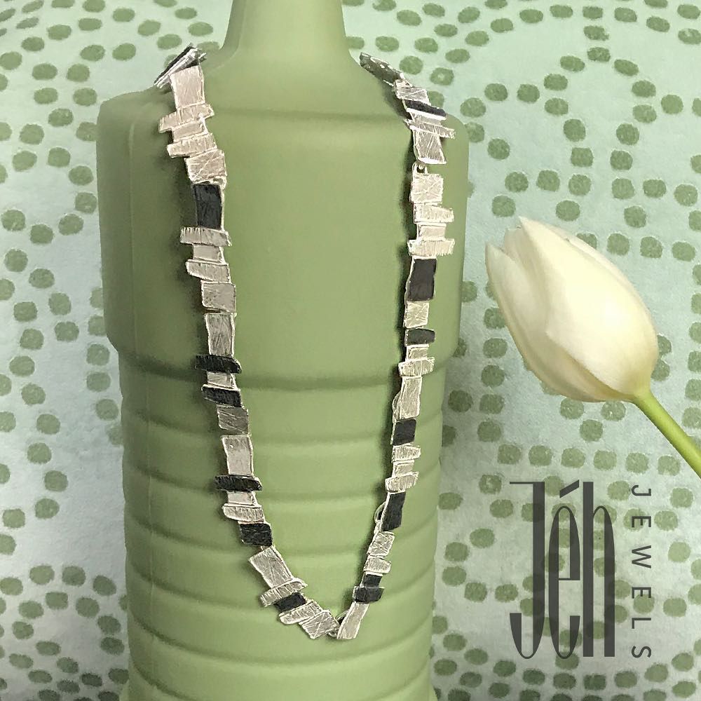 Zilveren en zwarte geometrische ketting gedrapeerd op een groene fles, witte tulp en stippenachtergrond.