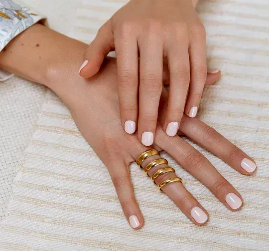 Close-up van handen met lichtroze nagellak en gouden ringen. Eén hand rust op de andere, op een wit, getextureerd oppervlak.