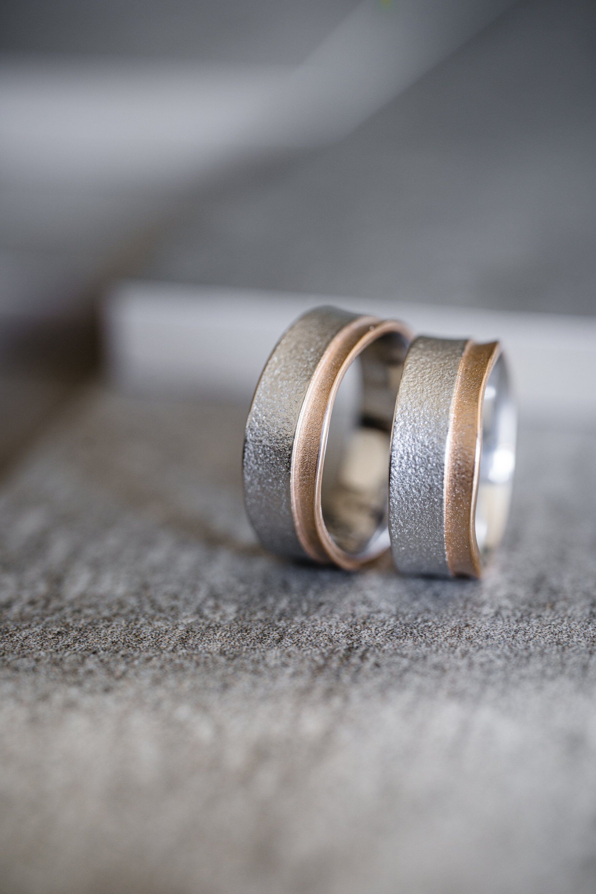 Twee trouwringen, zilver met een roségouden middenband, op een getextureerd grijs oppervlak.