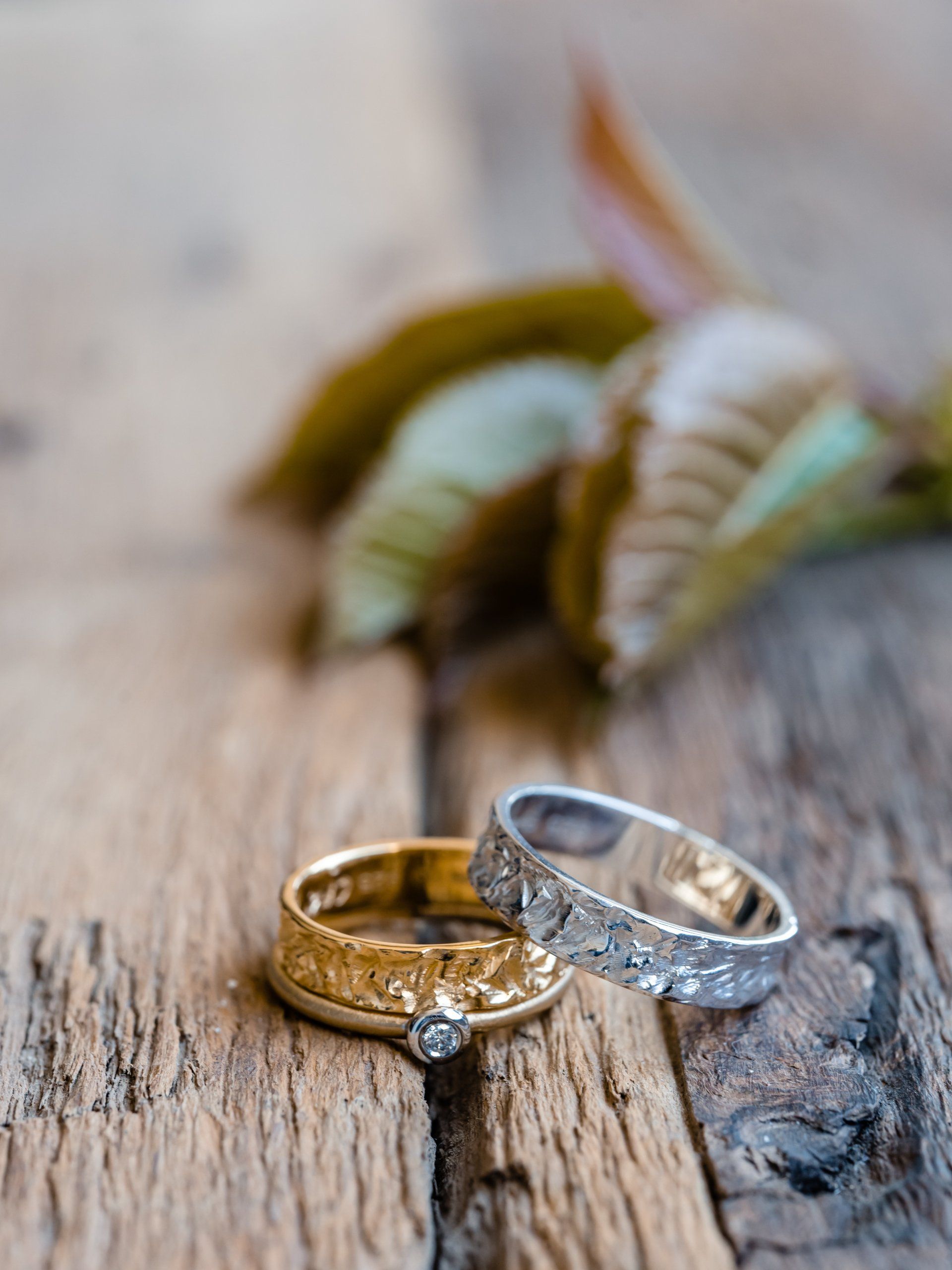 Twee trouwringen op rustiek hout, goud en zilver, met blad op de achtergrond.