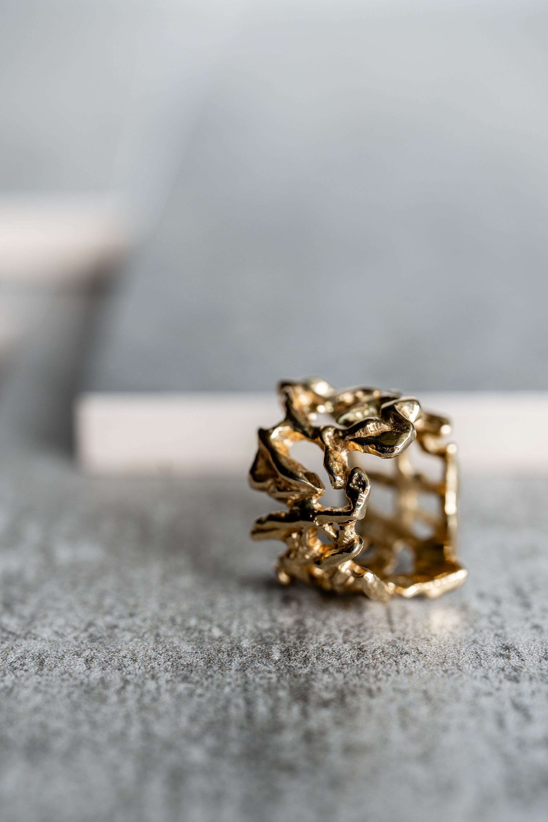 Goudkleurige, abstracte ring met een organisch, getextureerd design op een grijs oppervlak met onscherpe achtergrond.
