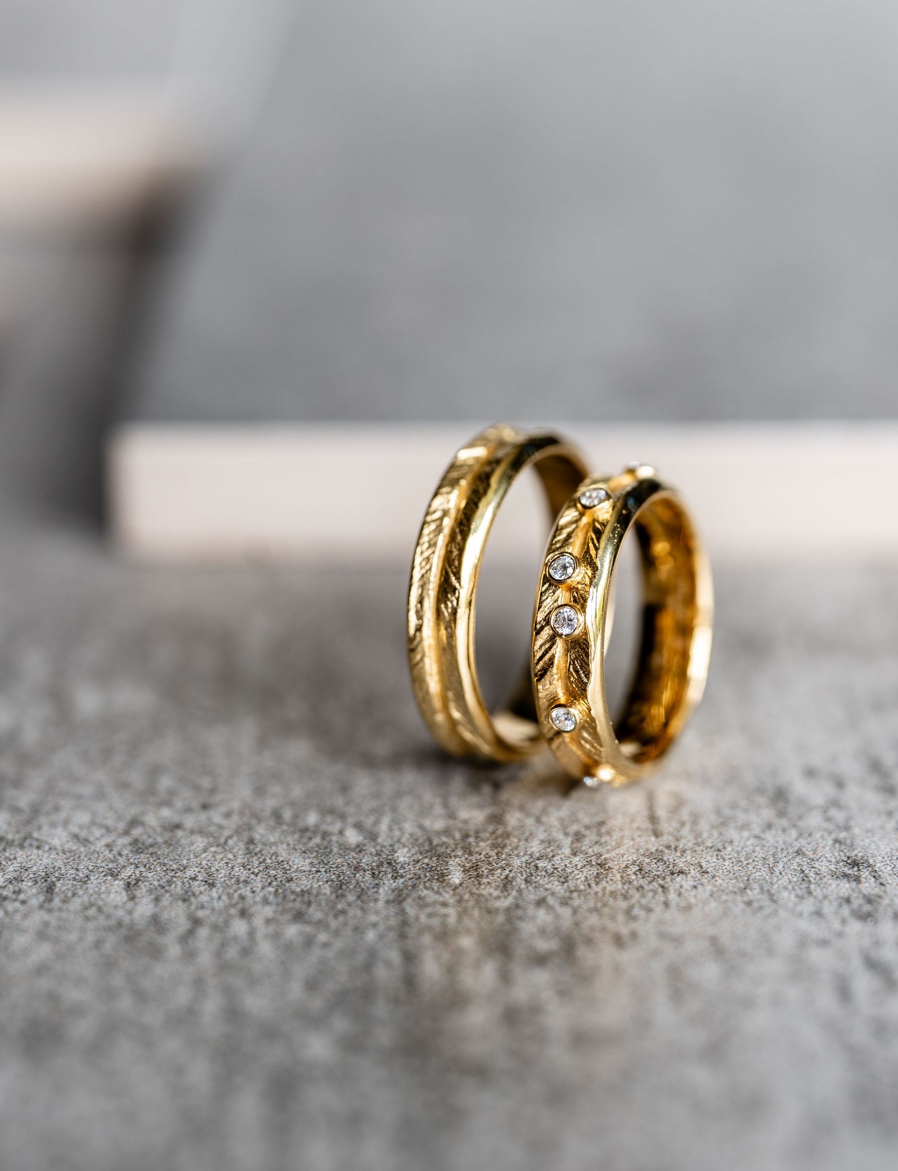 Twee gouden trouwringen op een getextureerd grijs oppervlak. De ringen hebben gedetailleerde ontwerpen, waarvan één met kleine steentjes.
