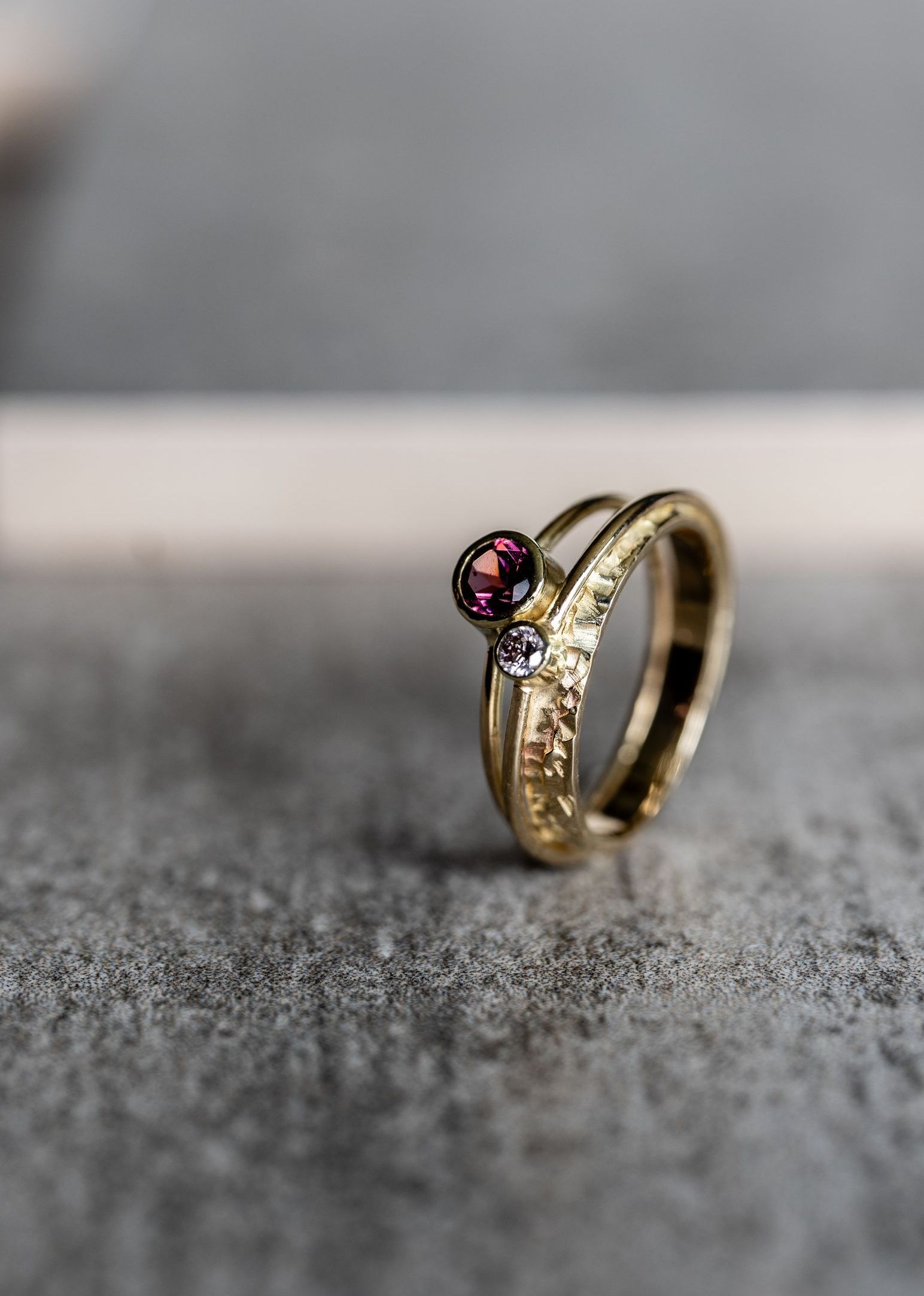 Gouden ring met een paarse edelsteen en kleinere witte stenen op een gestructureerde band, rustend op een grijs oppervlak.