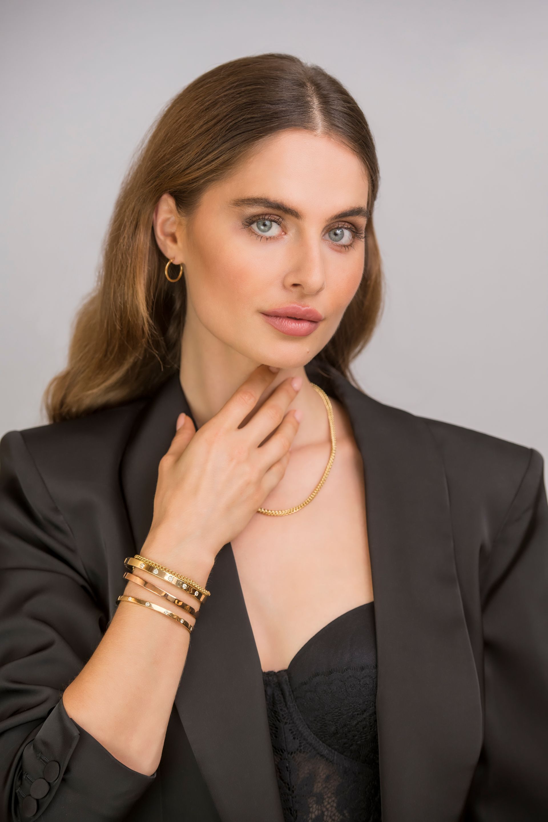 Vrouw met sieraden, zwart jasje en kanten top. Ze raakt haar nek aan en kijkt de toeschouwer aan.
