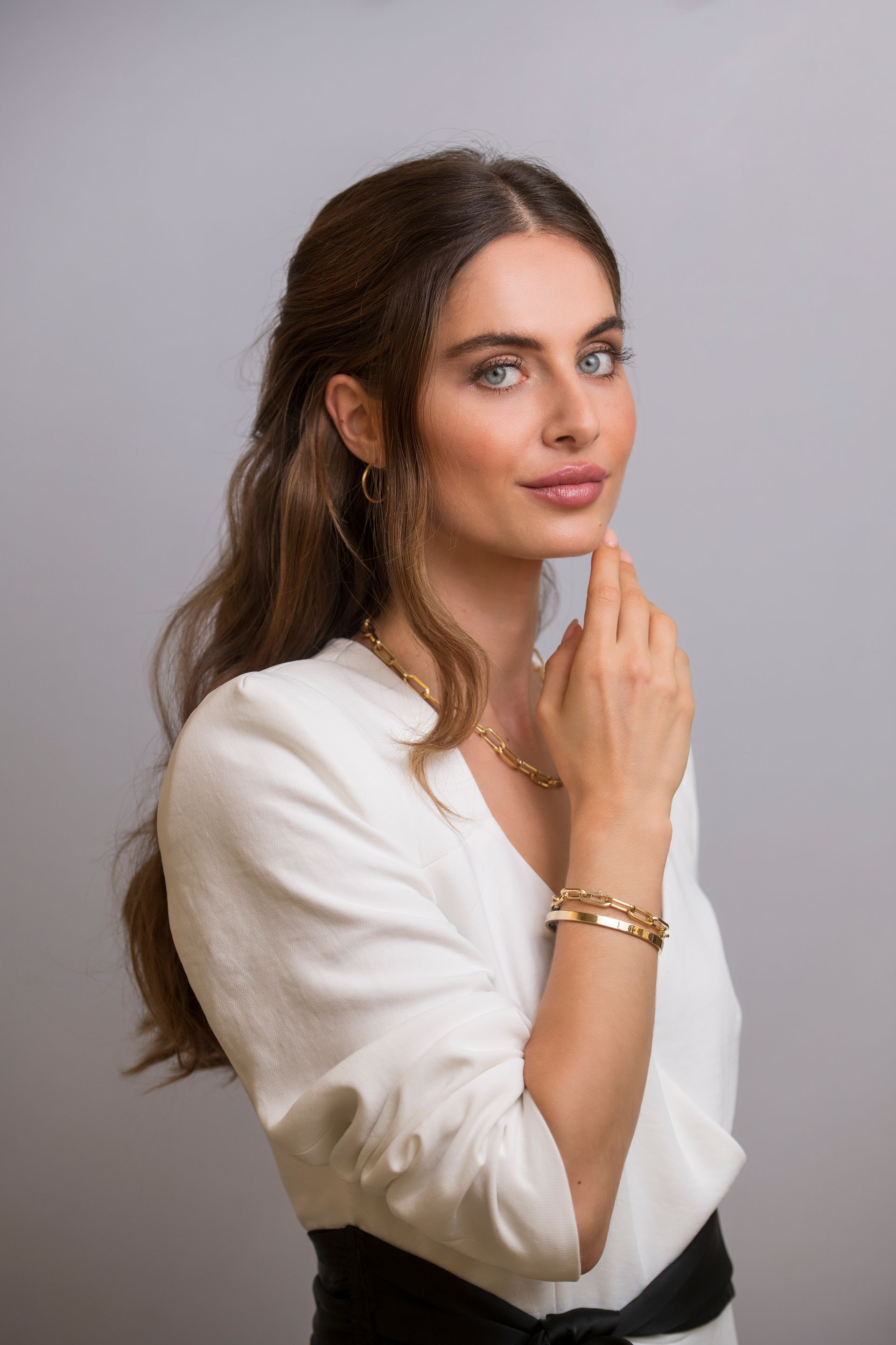 Vrouw in wit jasje, armband, hand bij haar gezicht, lichte achtergrond.