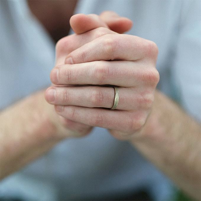 Handen ineengevouwen, met een trouwring om. Op de achtergrond zijn armen en een wit overhemd zichtbaar.