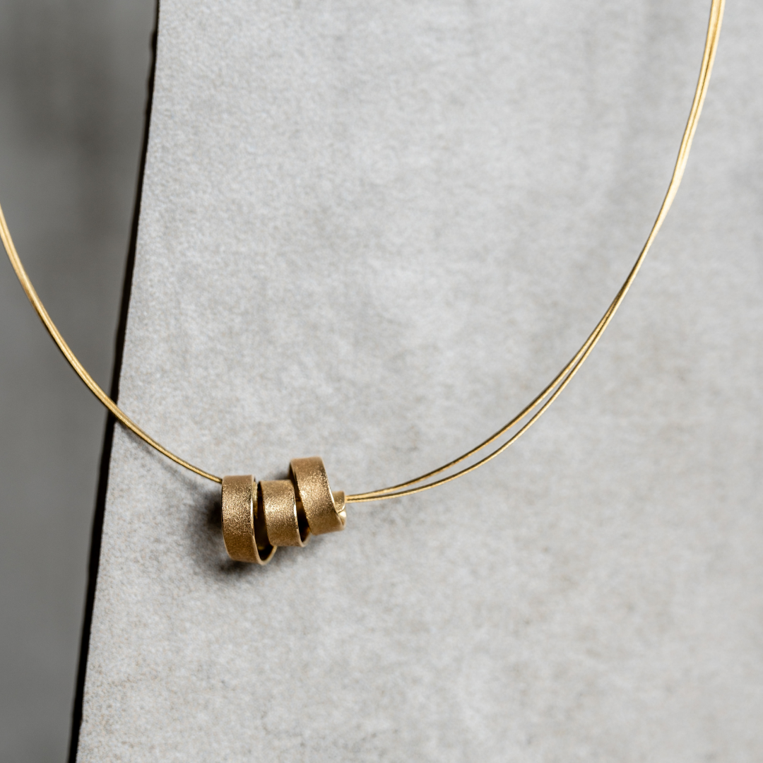 Gouden ketting met drie spiraalringen aan een dunne draad, tegen een grijze achtergrond.