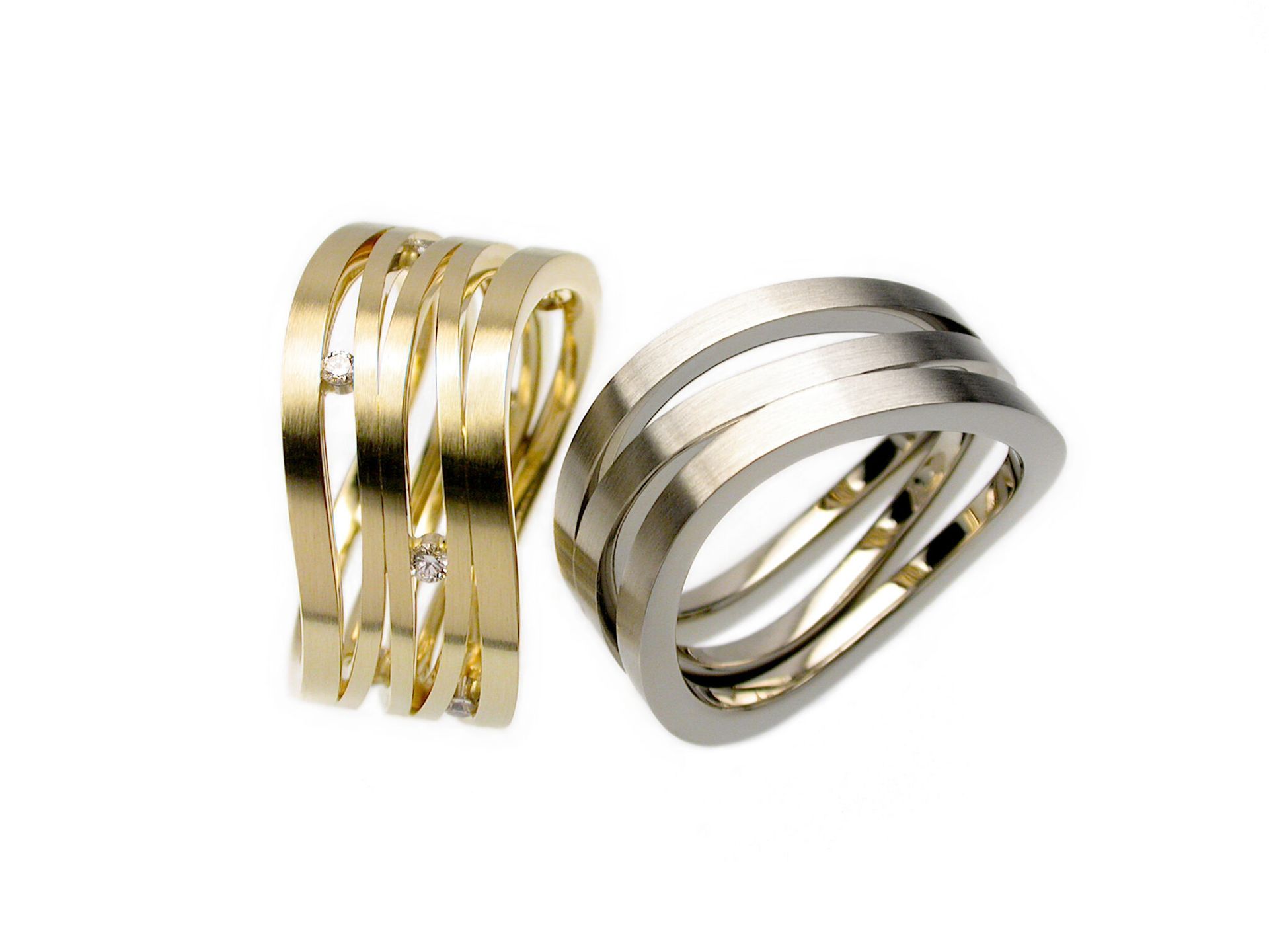 Twee golfvormige ringen, één van goud en één van zilver, elk met vier gebogen banden, waarvan één bezet met kleine diamanten in goud.