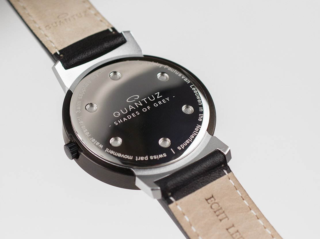 Achterkant van een horloge met een zwarte kast en leren band. De wijzerplaat is zilverkleurig en heeft het woord