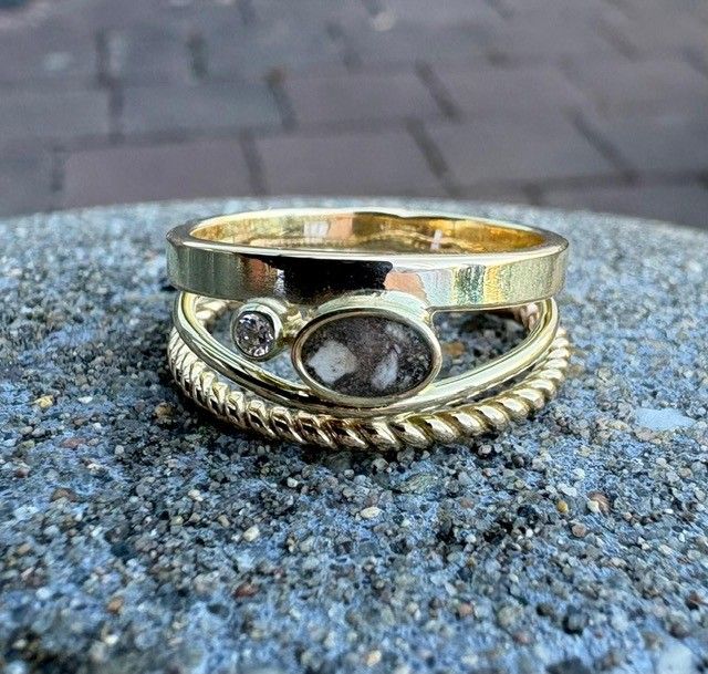 Gouden ringen op elkaar gestapeld, waarvan één met een ovaal inzetstuk, een diamant en een gedraaide band, op een gestructureerd oppervlak.