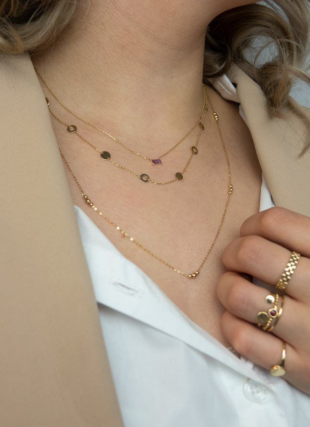 Vrouw draagt gouden kettingen en ringen, bruine blazer en wit overhemd.