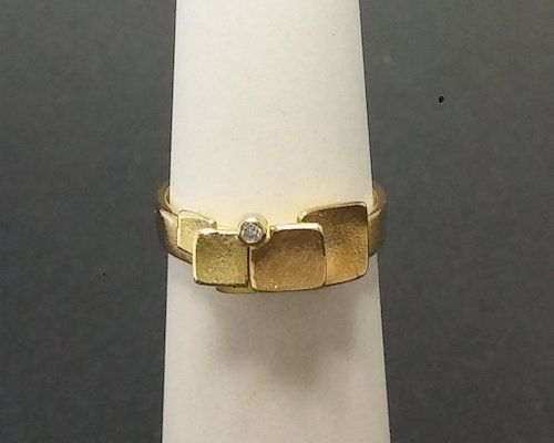 Gouden ring met getextureerde vierkante accenten en een centrale diamant op een witte standaard.
