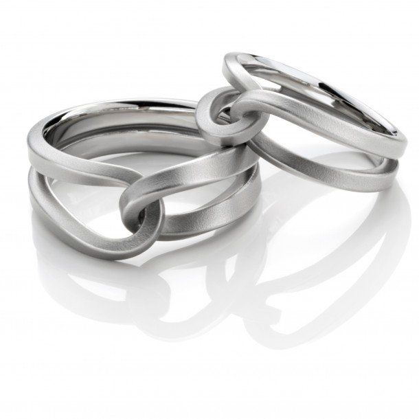 Twee in elkaar grijpende zilveren ringen, één met meerdere banden, de andere met een enkele band, op een wit oppervlak.
