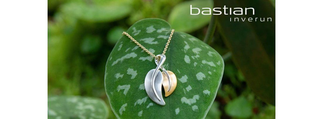 Een ketting met een hanger van zilver en goud op een groen blad; op de achtergrond het gebladerte.