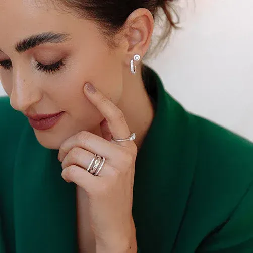 Vrouw in een groene blazer, met oorbellen en ringen, raakt haar gezicht aan en kijkt naar beneden.