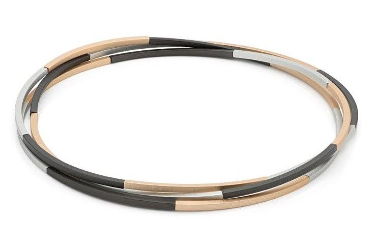 Armband met drie strengen: zwarte, zilveren en bruine metalen buisjes die in elkaar verstrengeld zijn, liggend op een witte achtergrond.