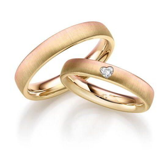 Twee gouden trouwringen, waarvan één met een hartvormige diamant, liggen naast elkaar op een wit oppervlak.