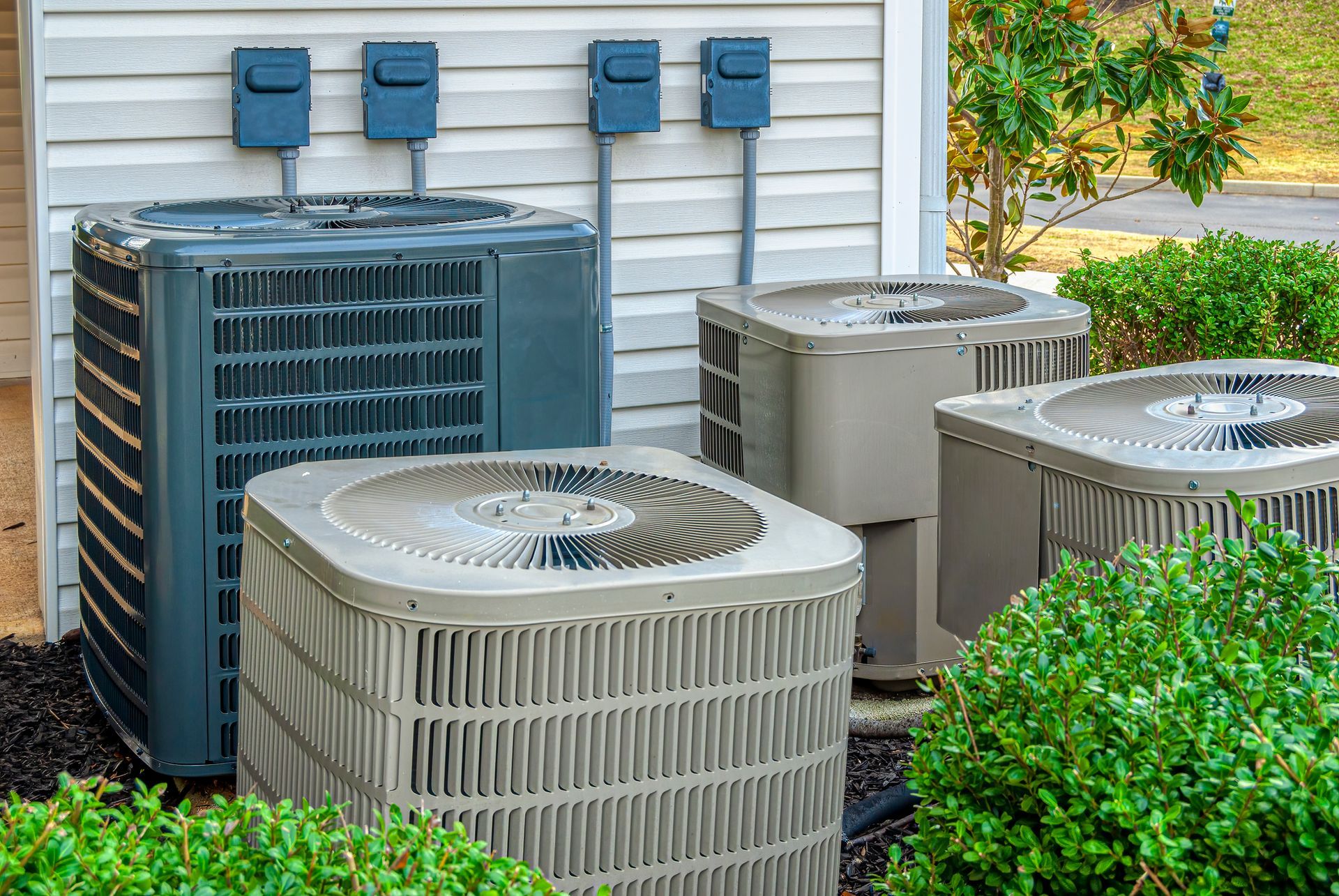 Outdoor HVAC units next to a building with electrical boxes and shrubbery.