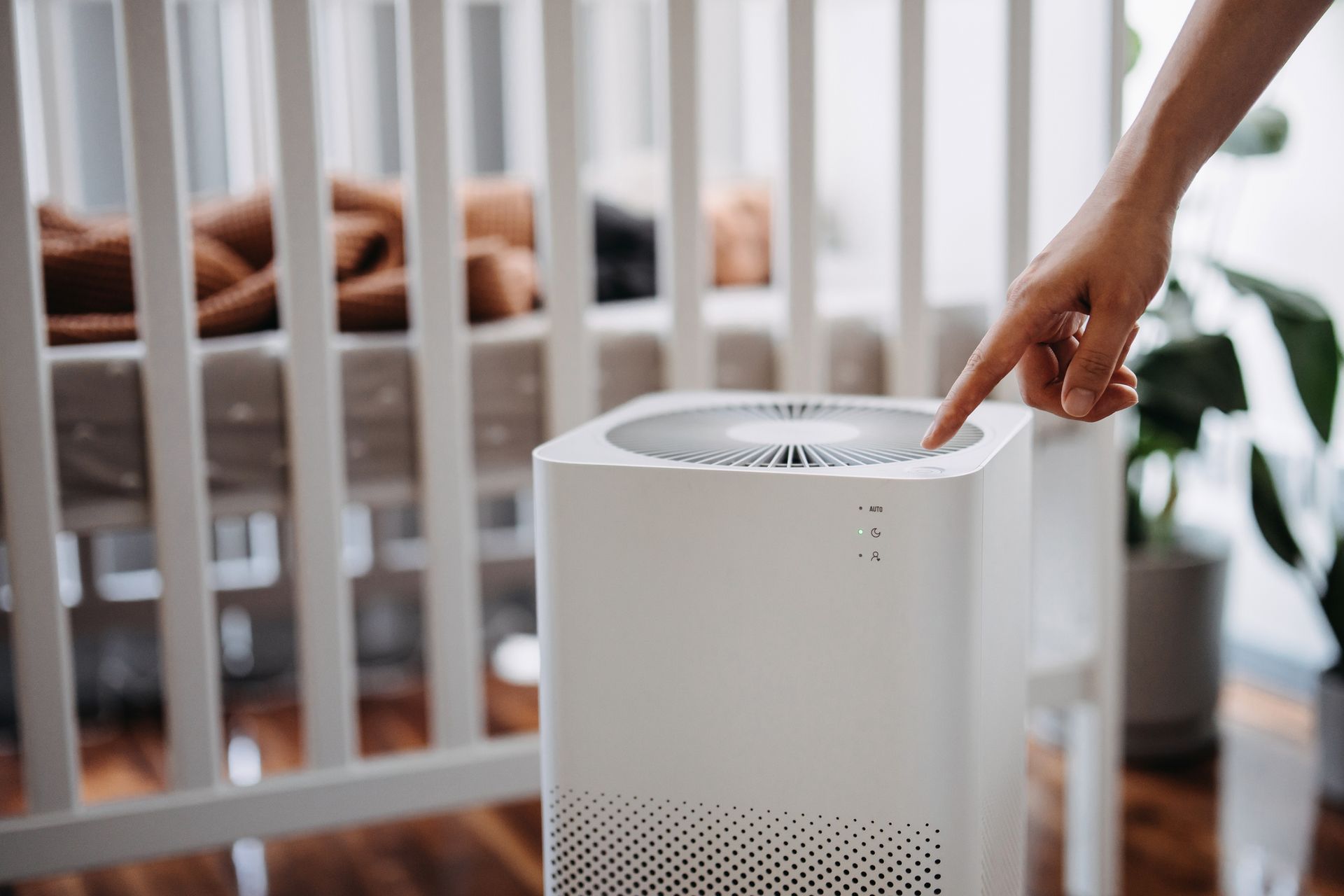 Hand presses air purifier button next to a baby crib. White purifier, white crib, indoor setting.
