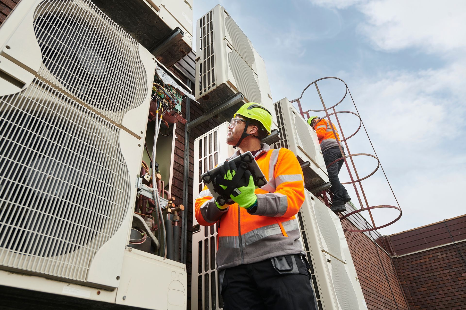 Two HVAC technicians inspecting units on a building exterior. One on a ladder, another holding a tablet.