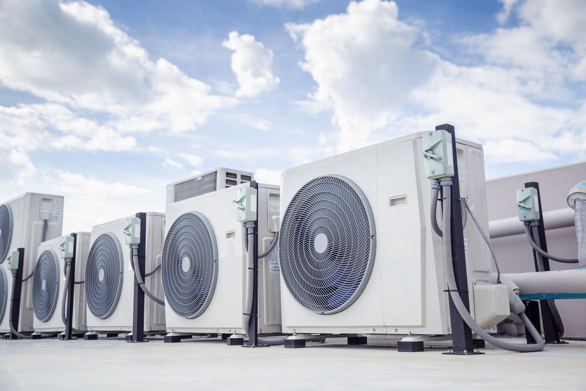 Row of white air conditioning units on a rooftop against a cloudy blue sky.