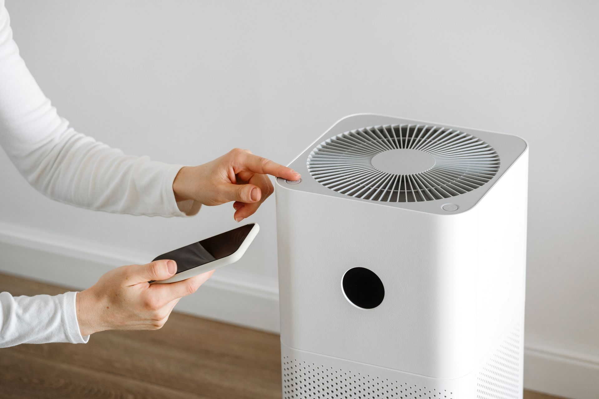 Person using a smartphone to operate a white air purifier in a room with a white wall.