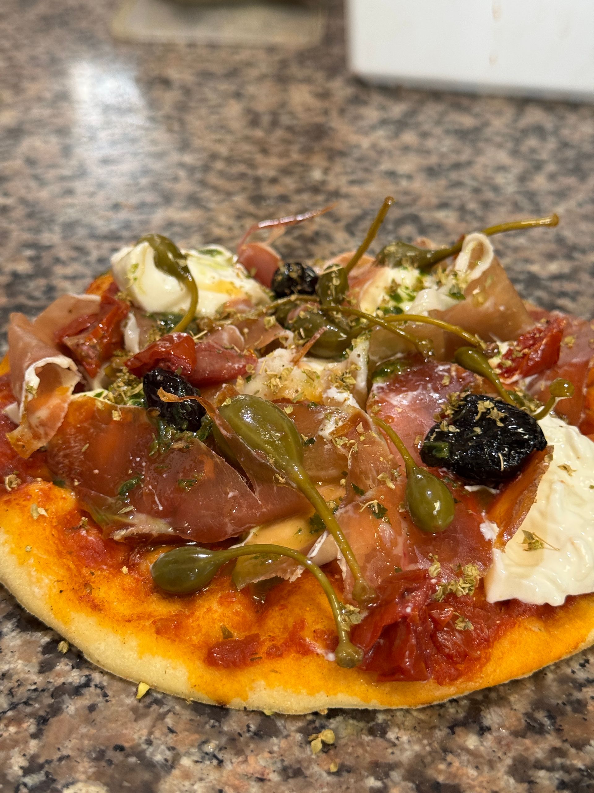 Petite pizza au prosciutto, câpres, olives et mozzarella sur une pâte mouchetée.
