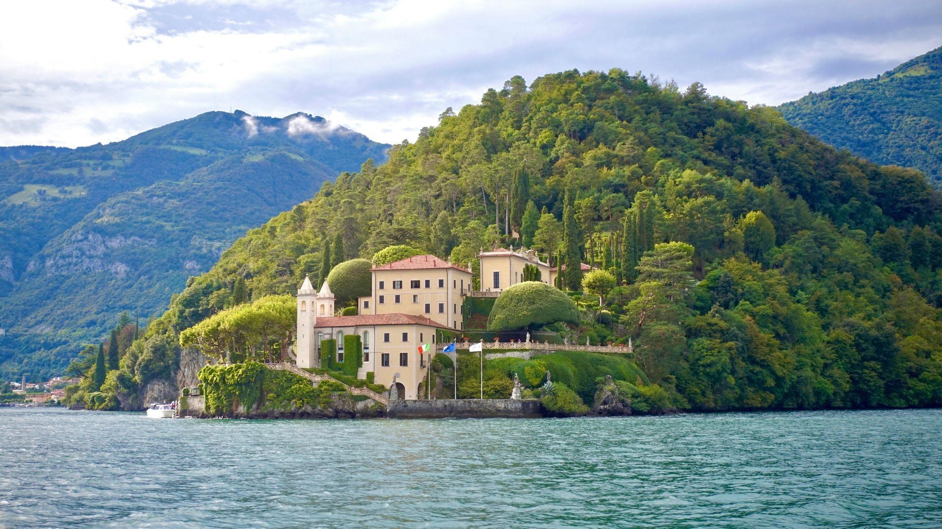 Villa beige aux toits de tuiles, perchée sur une colline verdoyante surplombant un lac, avec les montagnes en arrière-plan.