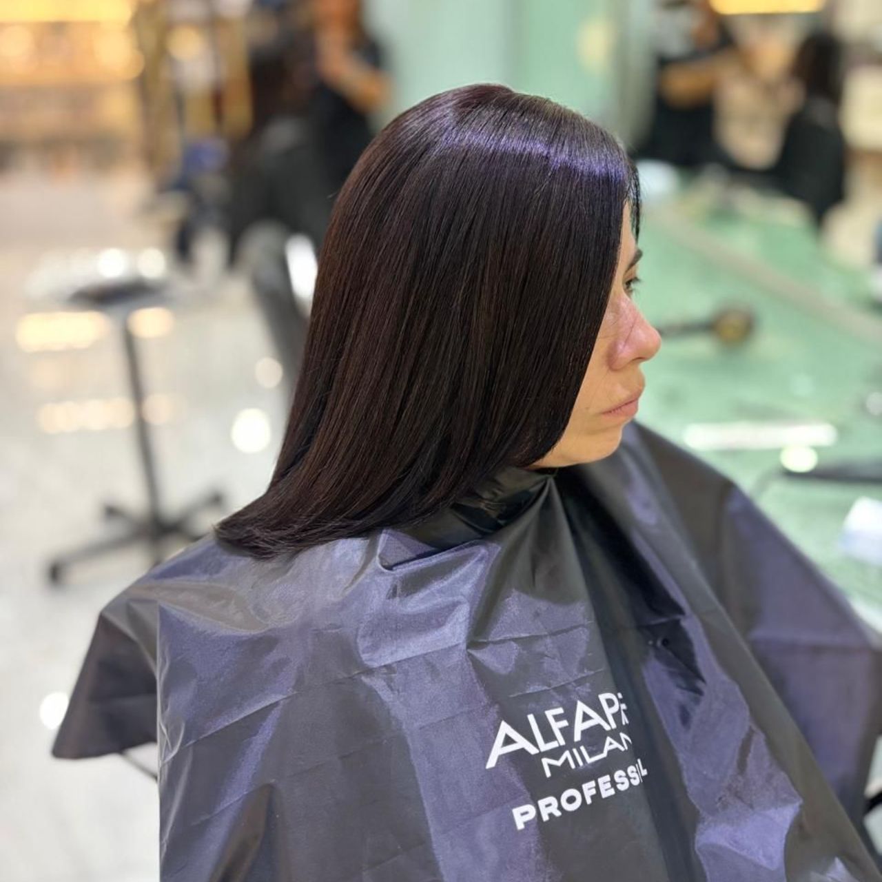Mujer con cabello largo y oscuro en una peluquería, con una capa negra, sentada de perfil.