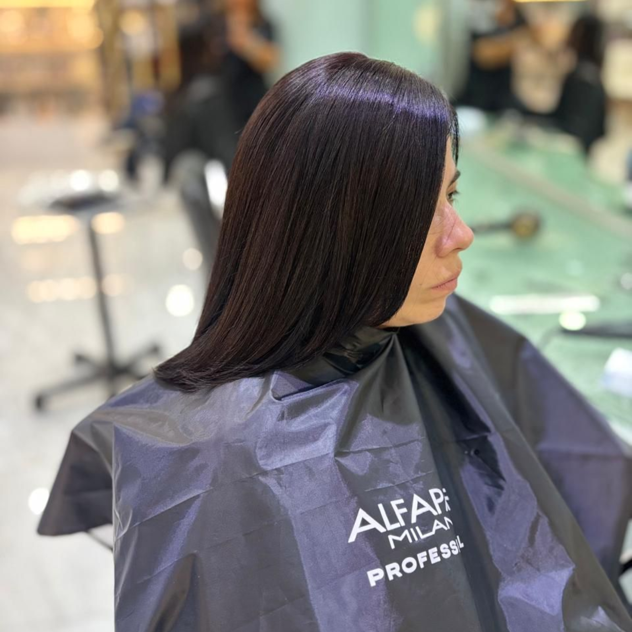 Mujer con cabello largo y oscuro con reflejos morados, sentada con una capa de peluquería en un salón de belleza.