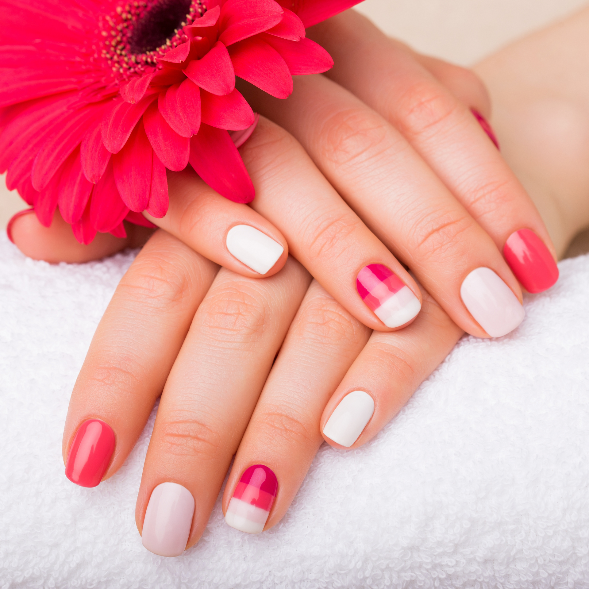 Manos con uñas pintadas de rosa y blanco junto a una flor rosa brillante sobre una toalla blanca.
