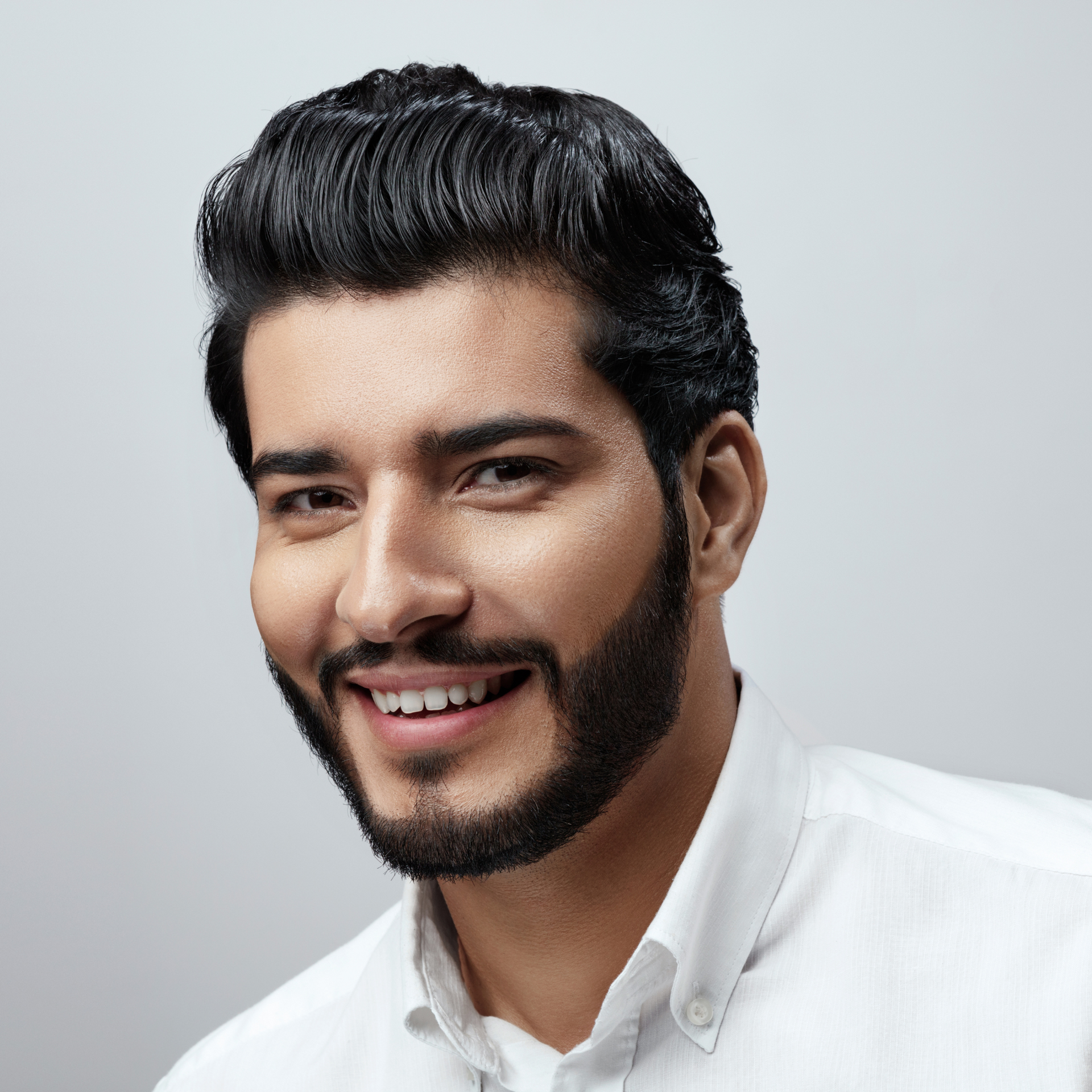 Hombre sonriente con cabello y barba oscuros, vestido con una camisa blanca, sobre un fondo gris claro.