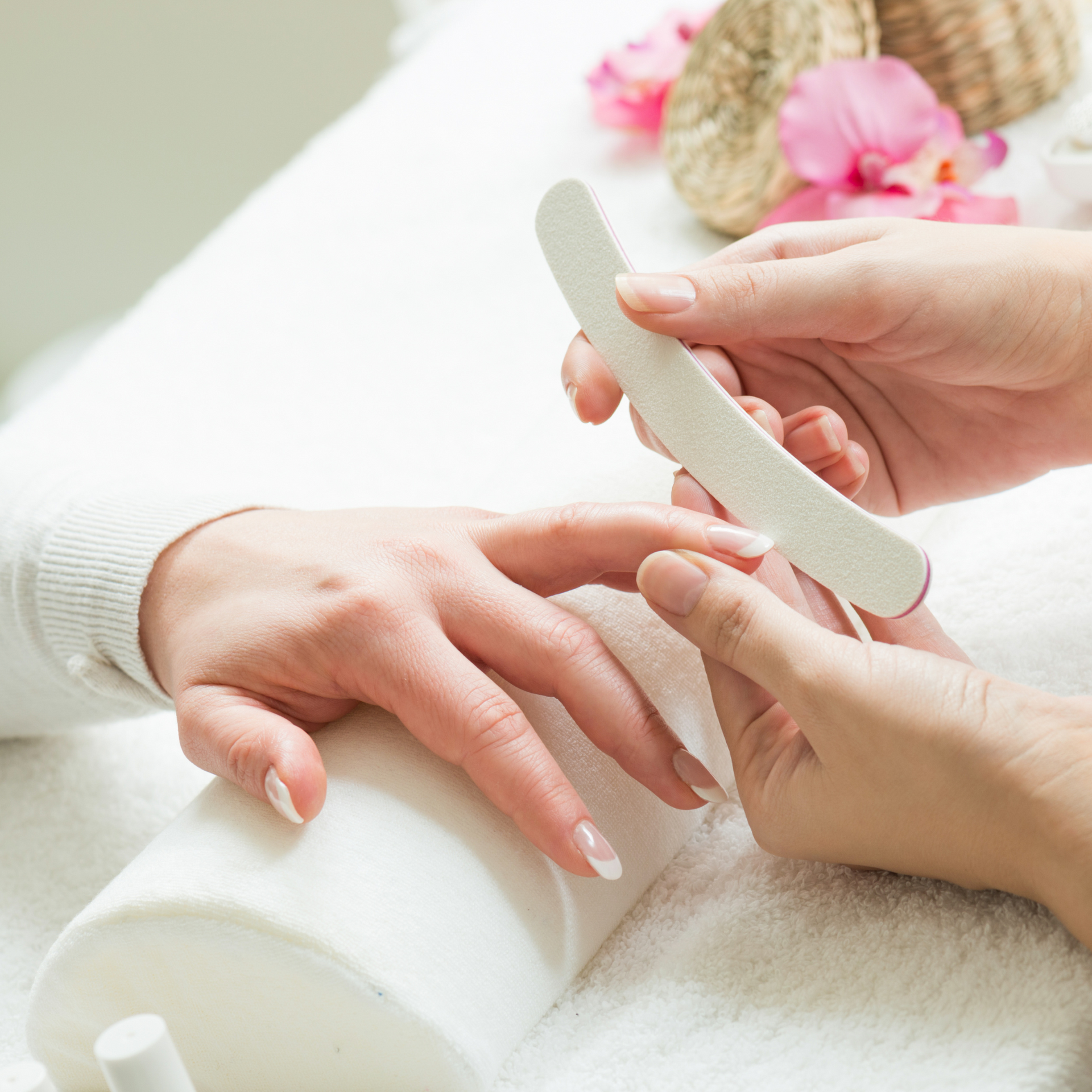 Manos limando las uñas de una persona en un luminoso spa con toallas blancas y flores rosas.