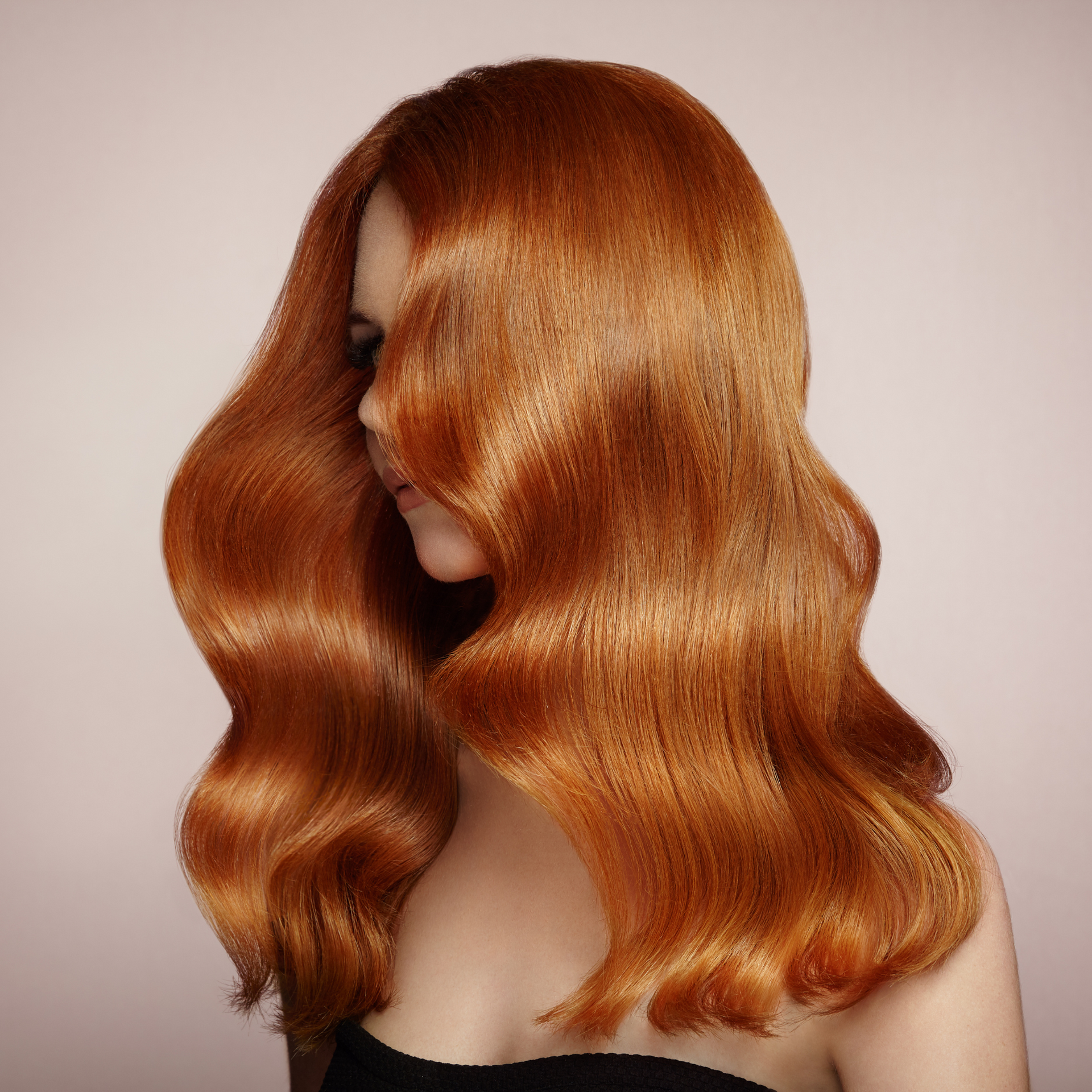 Cabello ondulado de color rojo cobrizo que cubre parcialmente un rostro sobre un fondo beige liso.